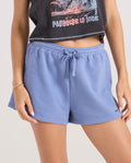 A person wears Volcom Women's Lived In Fleece Shorts in light blue with an elastic waistband and a black crop top with a "Paradise is Nice" graphic. Their hand, styled with a gold bracelet and light-colored nails, rests at their side.