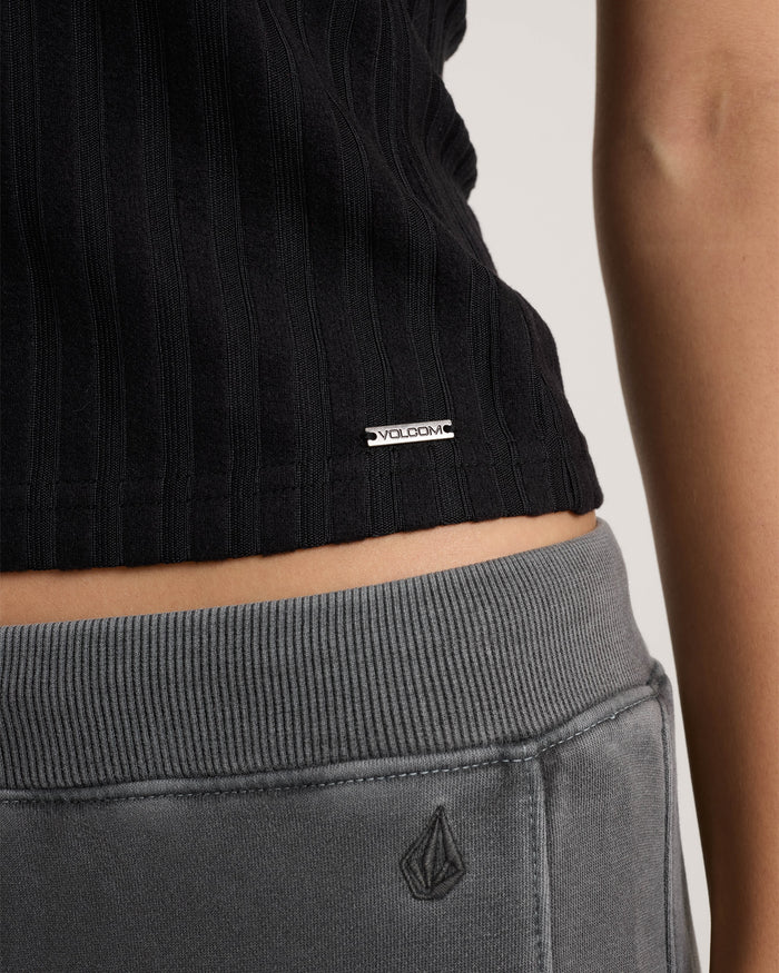 A close-up of a person wearing Volcom's Women's Lived In Rib Tank in black and gray Volcom sweatpants, focusing on the lower torso and hip area to highlight both brand logos.