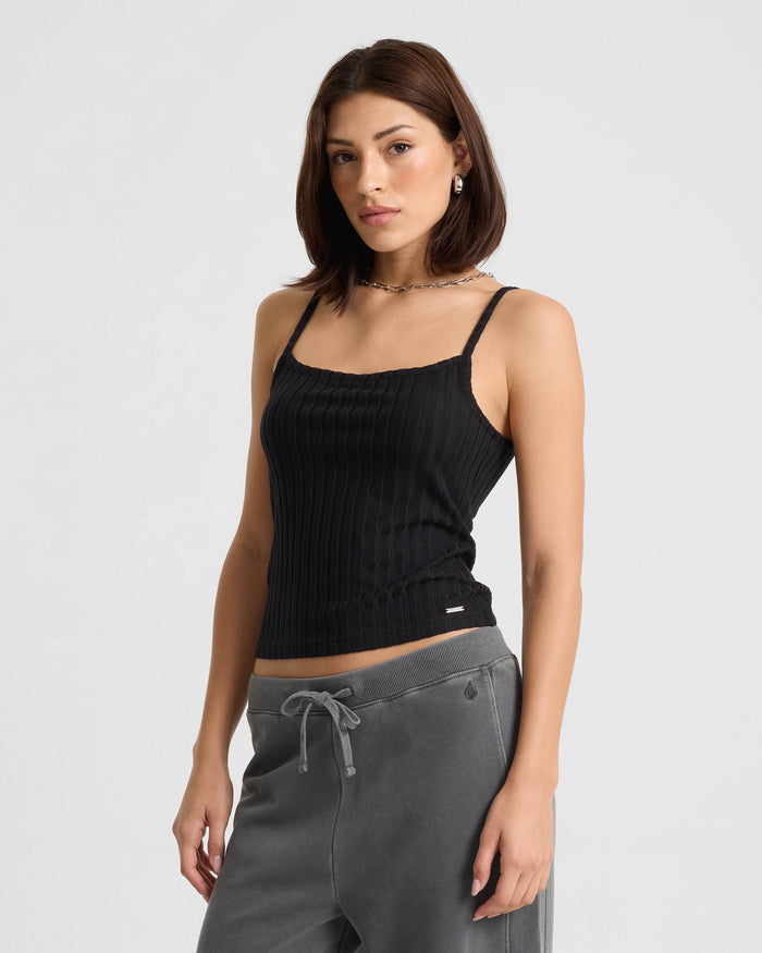 A woman with straight, shoulder-length brown hair wears the Volcom Women's Lived In Rib Tank in black and gray sweatpants, standing against a plain white background and looking at the camera with a neutral expression.