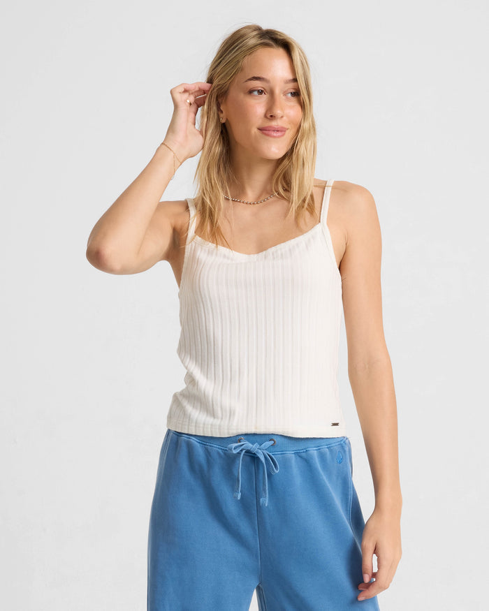 A blonde woman models the Volcom Women's Lived In Rib Tank in white with blue drawstring pants, posing against a light background and touching her hair as she looks to the side.