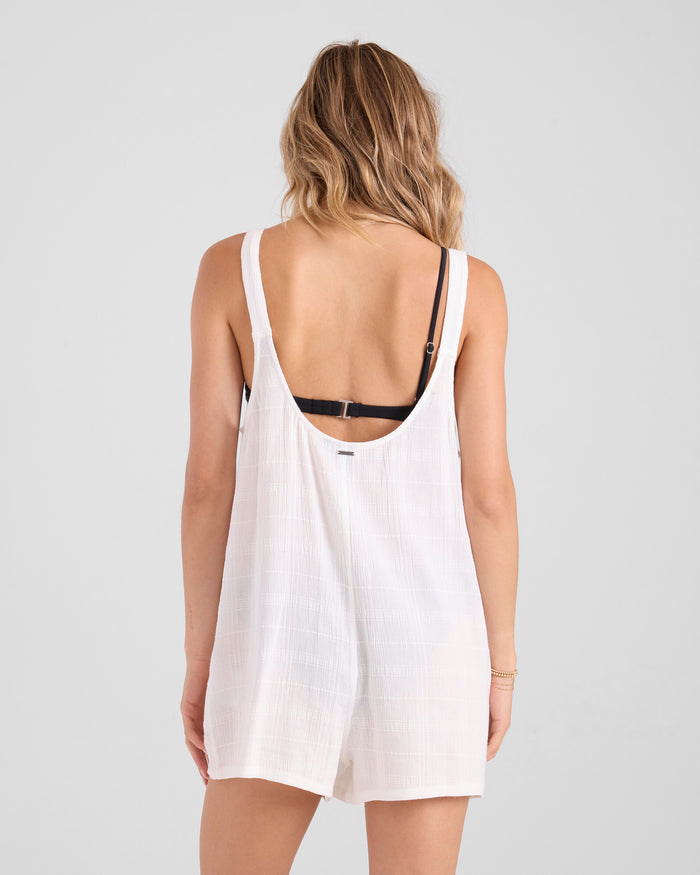 A woman with wavy, shoulder-length hair is seen from behind in the Volcom Women's Lived In Shorts Romper—a loose, white romper in a soft dobby weave styled over a black bra, with a low scooped back against a plain light background.