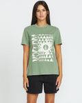 A woman with long dark hair wears the Volcom Women's Lock It Up Tee in sage green organic cotton and black shorts, standing against a plain white background.