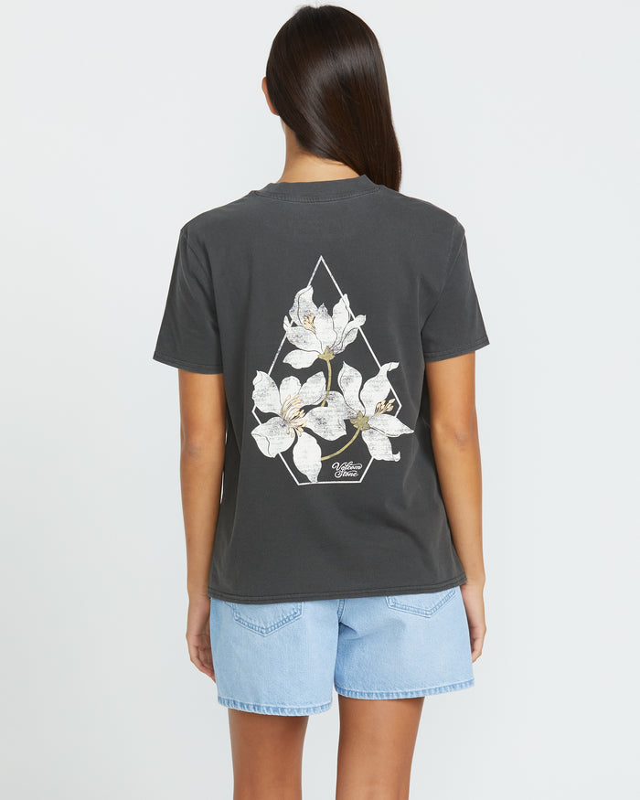 A woman faces away, showing the back of the Volcom Women's Lock It Up Tee in dark gray with white flower graphics, paired with light blue denim shorts against a plain white background for a casual, comfy look.