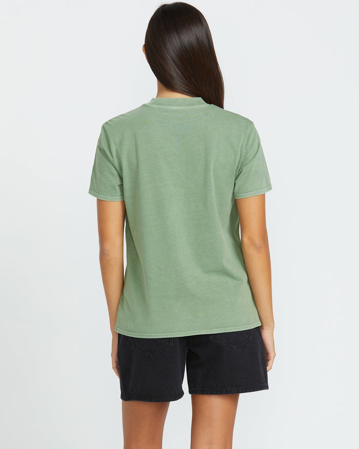 A woman with long dark hair, seen from behind, wears the Volcom Women's Lock It Up Tee in light green with black denim shorts. Her relaxed style stands out against the plain white background.