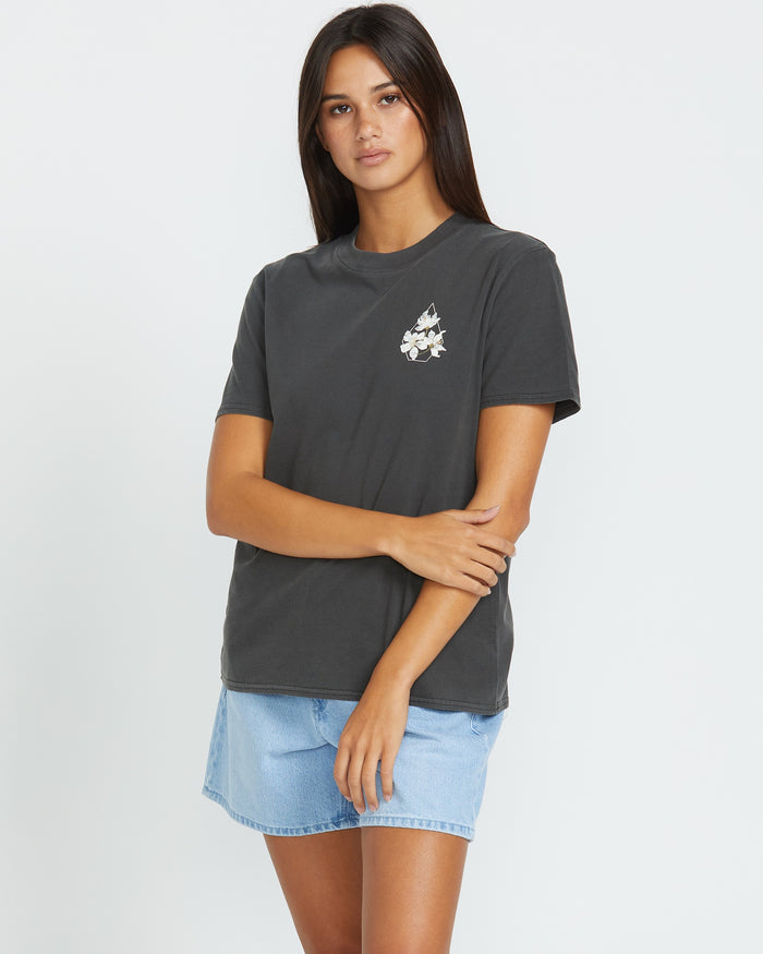 A woman with long dark hair wears the Volcom Women's Lock It Up Tee in charcoal, featuring a small floral graphic on the left chest, paired with a light blue denim skirt, standing against a plain white background.