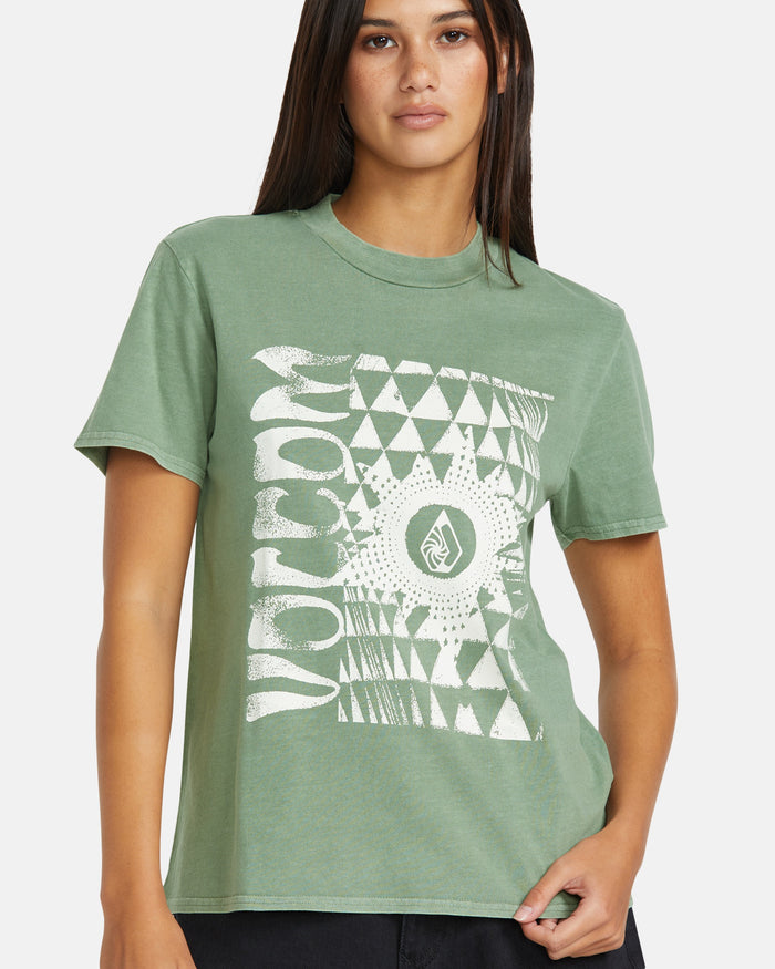 A woman models the Volcom Women’s Lock It Up Tee, a green relaxed-fit graphic tee made from organic cotton with a white abstract geometric design and "VOLCOM" on the front, standing against a plain background.
