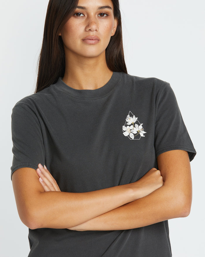 A woman with long dark hair stands with arms crossed, wearing Volcom's Women's Lock It Up Tee—an everyday dark gray organic cotton shirt featuring a small white floral design on the left chest.