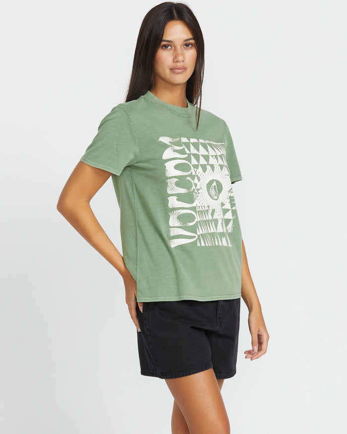 A woman with long dark hair poses against a white background wearing the Women's Lock It Up Tee by Volcom—an organic cotton green t-shirt with a white geometric graphic—and black shorts, standing with one hand on her hip.