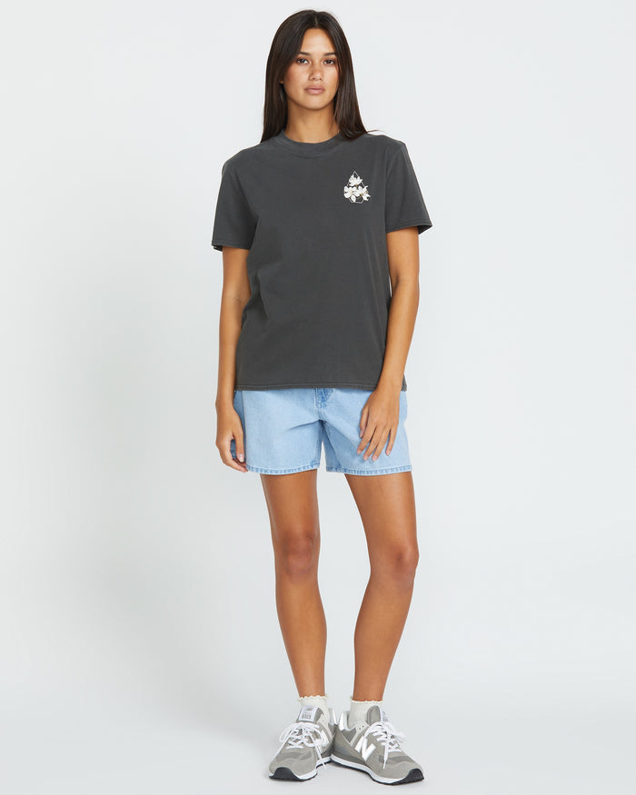 A woman stands facing forward wearing the Volcom Women's Lock It Up Tee in dark gray organic cotton with a small white graphic on the left chest, paired with light blue denim shorts and gray sneakers against a plain white background.