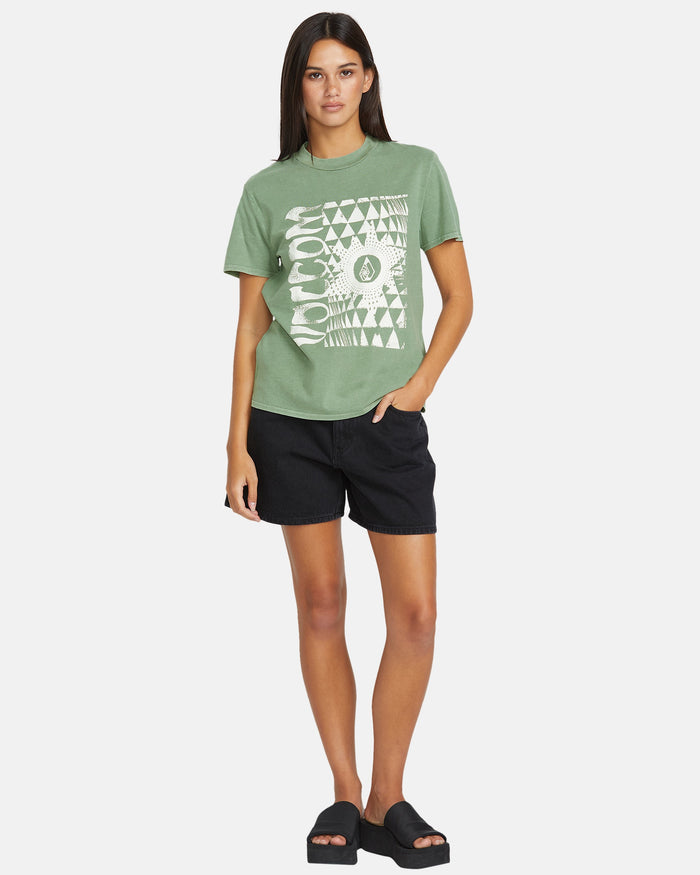 A woman faces forward in the Volcom Women's Lock It Up Tee, styled with black shorts and slide sandals. She has straight brown hair, a neutral look, and one hand in her pocket against a plain white background.