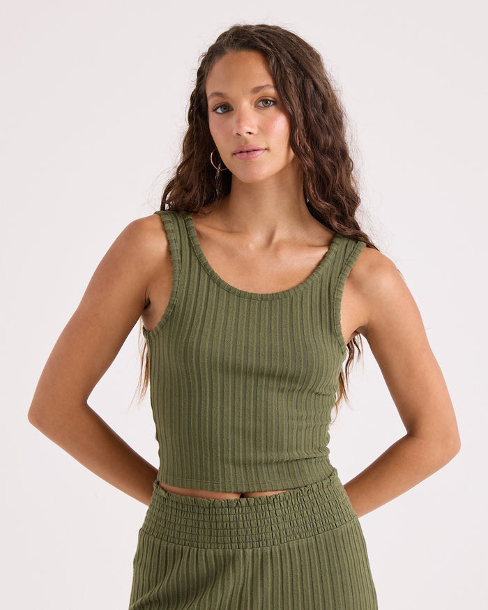 A woman with long wavy hair wears the Volcom Women's Lounge Bra Tank in olive green, featuring a flattering scoop neck and ribbed texture, paired with matching high-waisted bottoms. She stands against a plain light background with her hands resting behind her back.