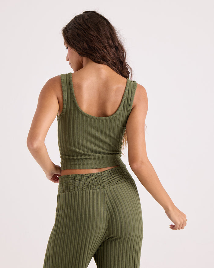 A woman with long brown hair stands facing away, her left arm slightly raised, showcasing the comfortable, stretchy knit fabric of the Volcom Women's Lounge Bra Tank in olive green with a ribbed texture, scoop neck, and matching high-waisted pants.