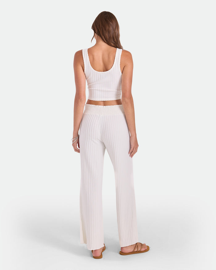 A woman stands facing away, wearing the Volcom Women's Lounge Bra Tank—an everyday essential. She pairs it with high-waisted, wide-leg pants and brown sandals. Her long hair flows down her back against a plain white background.