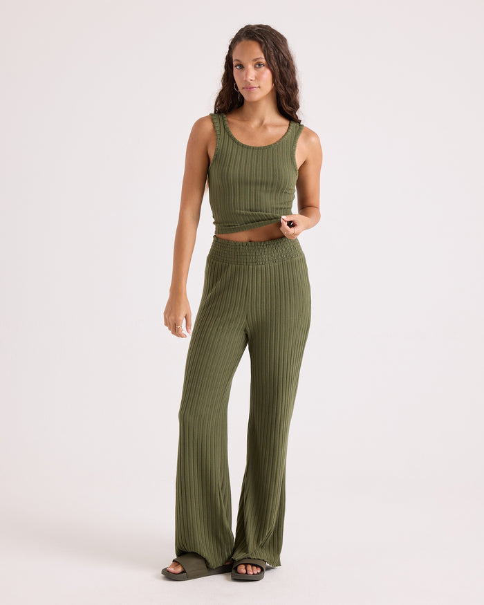 A woman stands against a plain background wearing the Volcom Women's Lounge Bra Tank, a sleeveless olive green top with a scoop neckline crafted from stretchy ribbed knit fabric, paired with matching high-waisted wide-leg ribbed pants and black sandals.