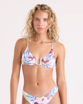 A woman with long curly blonde hair models the Volcom Women's Lush Jungle Uwire Bralette, a floral bikini featuring pink, blue, and green patterns with adjustable straps and a U-wire bralette top made from recycled nylon. She poses against a plain light background.