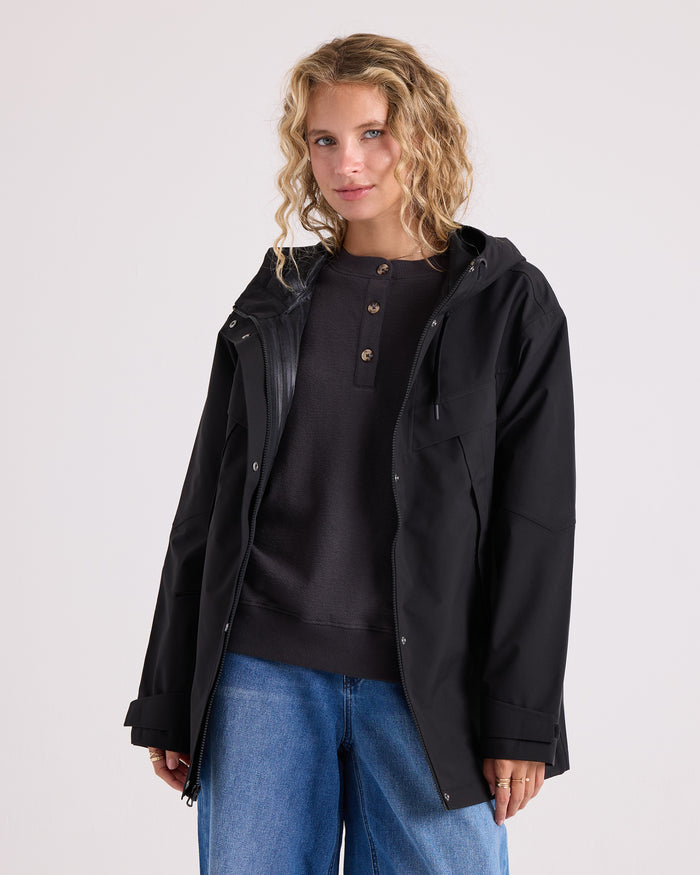 A woman with curly blonde hair wears the Volcom Women’s Margot Rain Jacket in black over a dark henley shirt and blue jeans, standing against a plain light background and looking at the camera with a slight smile.