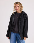 A woman with curly blonde hair wears the Volcom Women’s Margot Rain Jacket in black over a dark henley shirt and blue jeans, standing against a plain light background and looking at the camera with a slight smile.