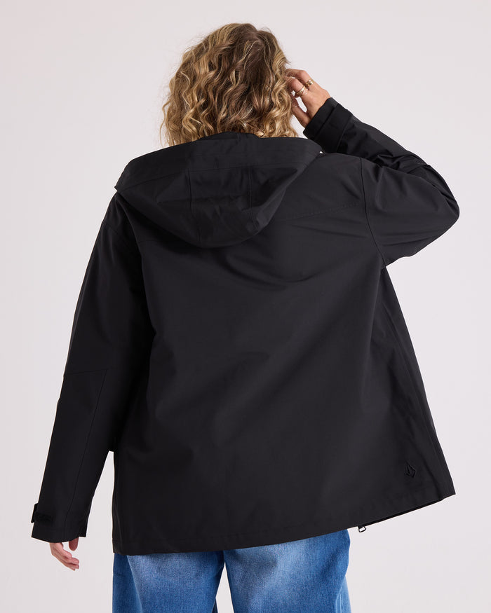 A person with wavy blonde hair, wearing the Volcom Women’s Margot Rain Jacket in black and blue jeans, stands facing away from the camera, with one hand touching their hair.