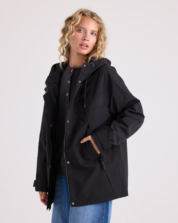 A person with wavy blonde hair wears the Volcom Women’s Margot Rain Jacket—a black, water resistant hooded jacket—over a dark shirt and blue jeans, standing against a plain light background with one hand in their jacket pocket.