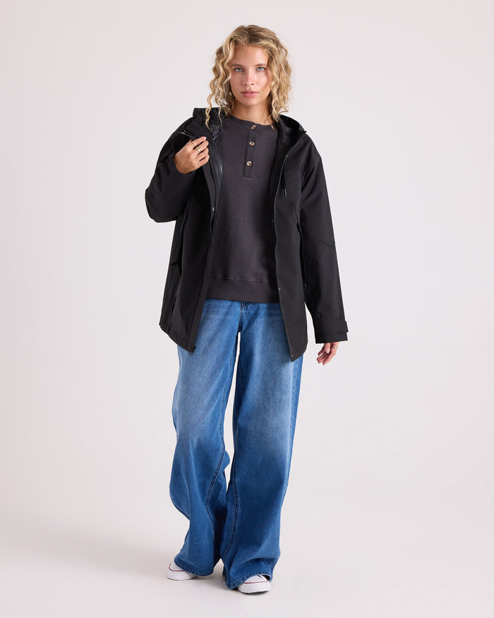 A person with wavy blonde hair wears the Volcom Women’s Margot Rain Jacket, which is water resistant and black, layered over a dark henley shirt and paired with loose blue jeans, standing against a plain light background.