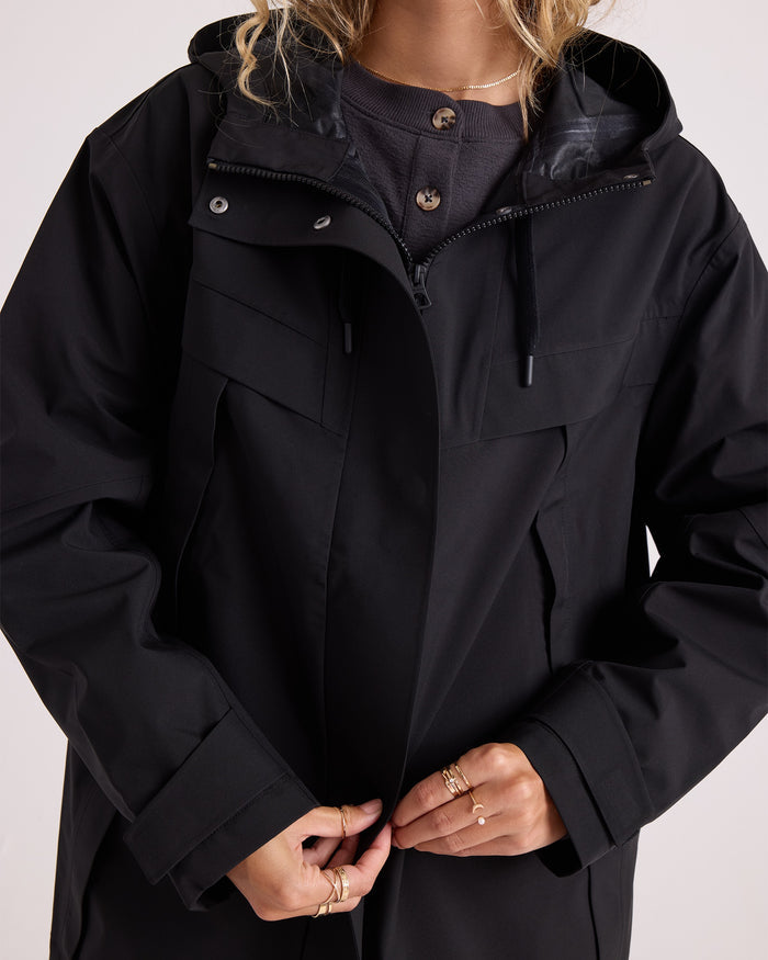 A person with wavy blonde hair wears the Volcom Women’s Margot Rain Jacket—a black, water-resistant, hooded style—over a dark top, fastening it at the front. Only the lower half of their face is visible.
