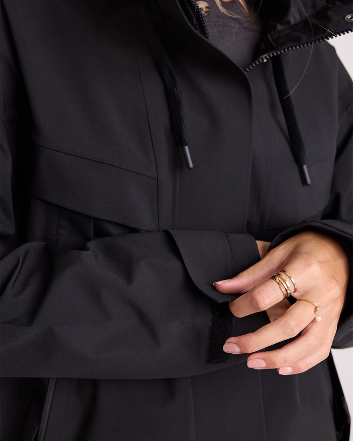 Close-up of a person adjusting the sleeve of the Volcom Women’s Margot Rain Jacket in black, displaying their hand adorned with several gold rings.