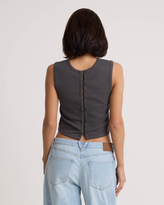 A person with short dark hair, seen from behind, wears the Volcom Women's Mary Mack Tank in Vintage Black—a cotton blend sleeveless crew-neck top featuring buttons down the back—paired with light blue jeans against a plain light background.