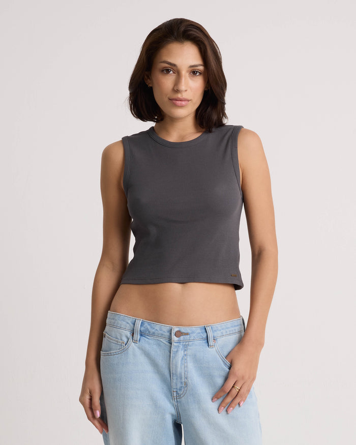 A woman with shoulder-length brown hair wears the Volcom Women's Mary Mack Tank in Vintage Black, featuring a fitted crew-neck design crafted from a soft cotton blend, paired with light blue jeans. She stands against a plain light background with a neutral expression.