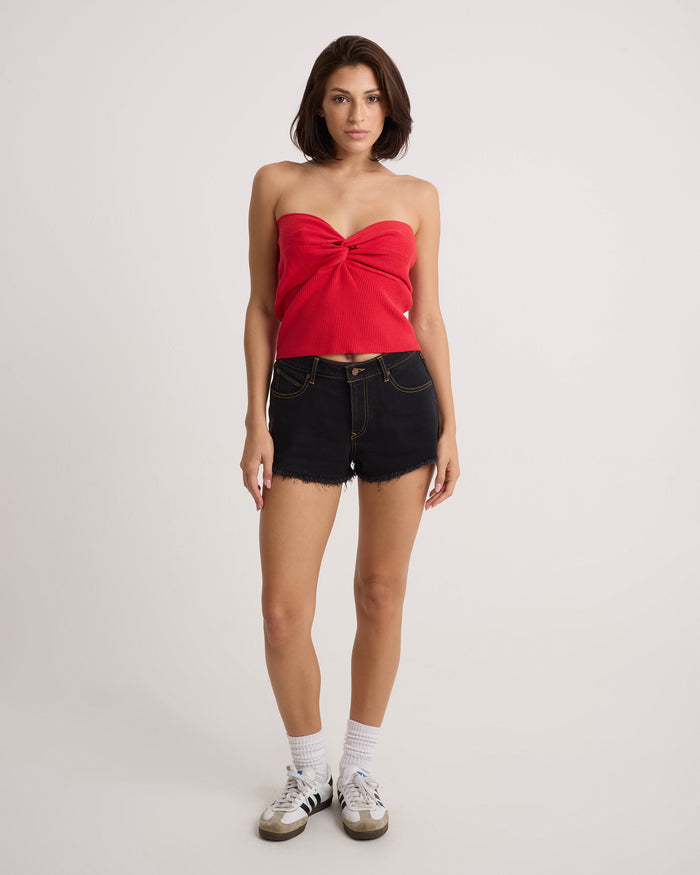 A woman faces forward against a plain light background, wearing the Volcom Women's Merlin Twist Top in persimmon, black denim shorts, white socks, and white sneakers with black stripes.