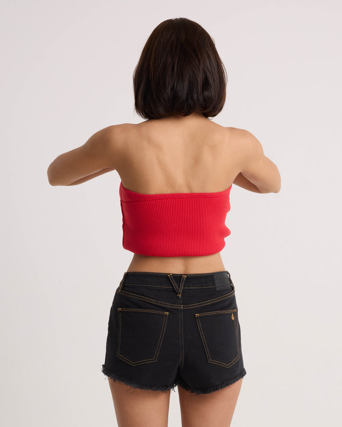 A woman with short dark hair is seen from behind, wearing a Volcom Women's Merlin Twist Top in persimmon and black denim shorts, standing against a plain light background with her arms slightly raised.