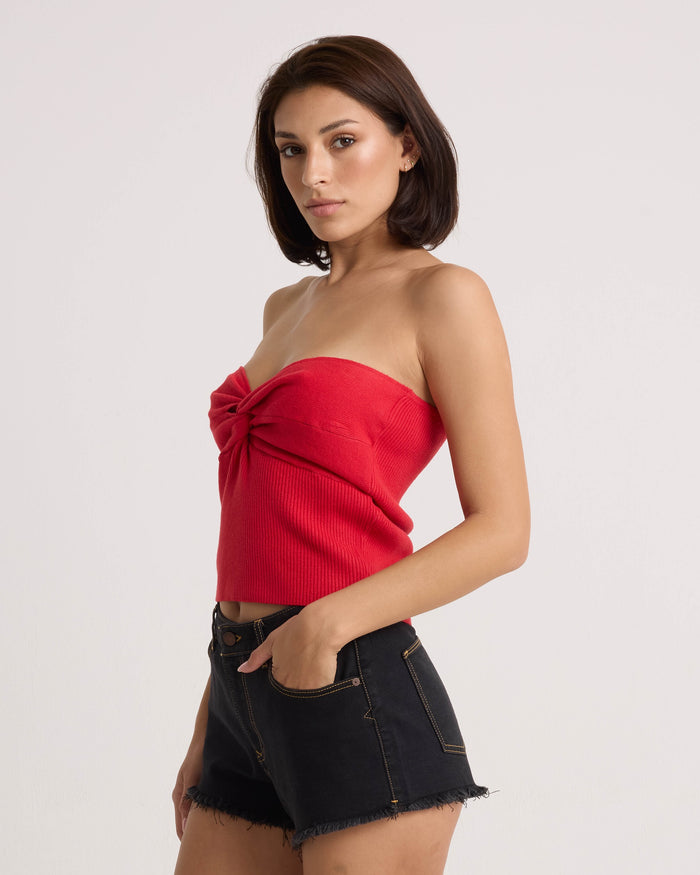 A woman with straight brown hair stands sideways against a white background, wearing the Volcom Women's Merlin Twist Top in persimmon and black frayed-hem denim shorts.