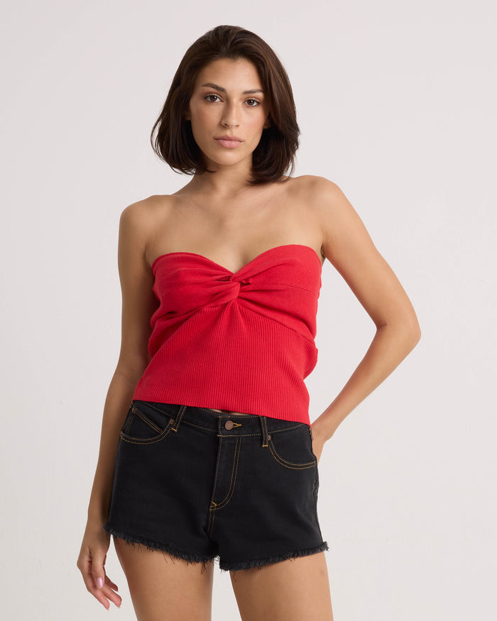 A woman with straight brown hair wears the Volcom Women's Merlin Twist Top in persimmon and black denim shorts, standing against a plain light background with one hand in her pocket and a neutral expression.