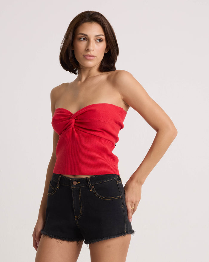 A woman with shoulder-length brown hair models the Volcom Women's Merlin Twist Top in persimmon and black denim shorts, standing against a plain light background, gazing to the side with one hand on her hip.