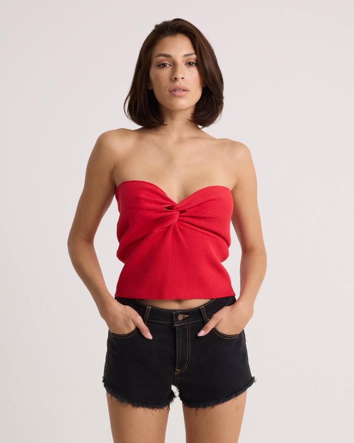 A woman with straight brown hair models the Volcom Women's Merlin Twist Top in persimmon with black denim shorts. She stands against a plain white background, hands in her pockets, and looks at the camera.