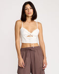 A woman with straight dark hair wears the Volcom Women's Money Mama Cami—a fitted cami top with a front tie, cutout detail, and smocked back—paired with brown high-waisted cargo pants, standing against a plain light background.