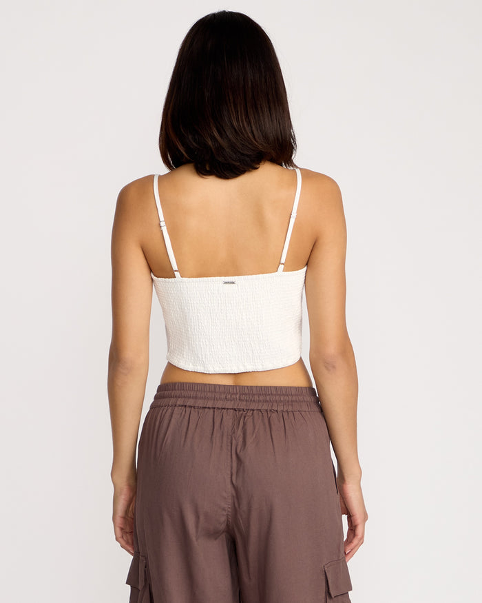 A woman stands facing away, wearing the Volcom Women's Money Mama Cami in white, featuring thin straps and a smocked back, paired with high-waisted brown pants with pockets, against a plain white background.