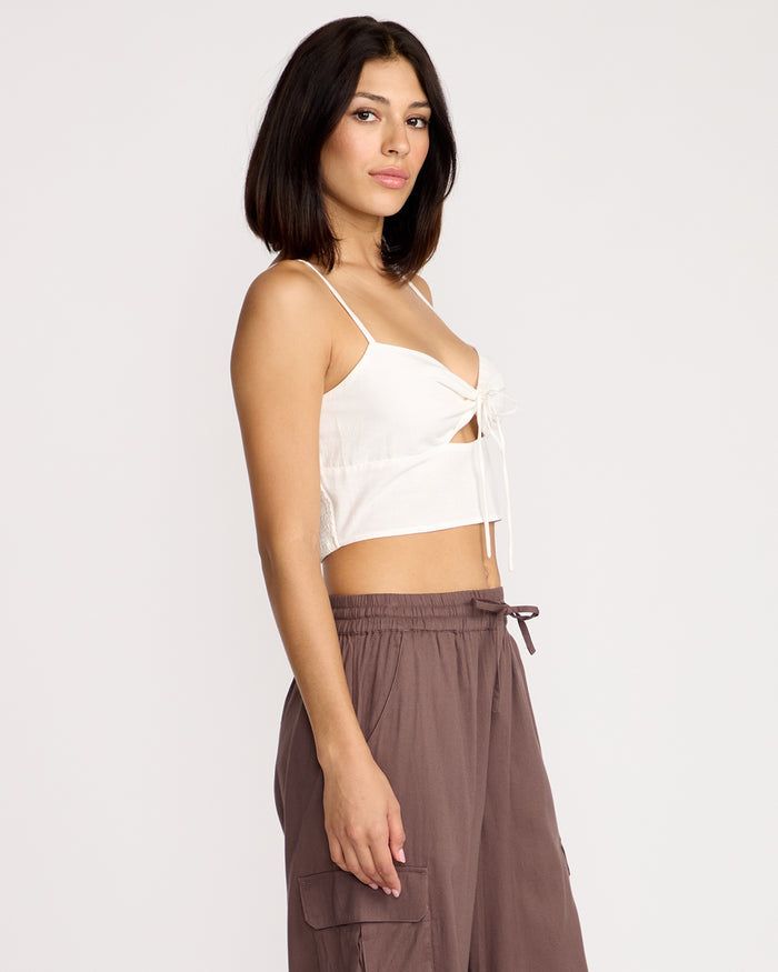 A woman stands sideways, confidently facing the camera against a plain light background, wearing the Volcom Women's Money Mama Cami—a white, fitted sleeveless top featuring a front tie and smocked back—paired with loose brown cargo pants.