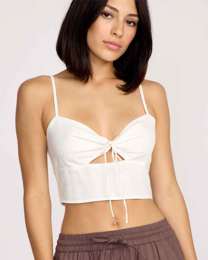 A woman with straight dark hair wears the Volcom Women's Money Mama Cami—a white fitted top featuring a front tie and smocked back—paired with brown pants, standing against a plain light background.
