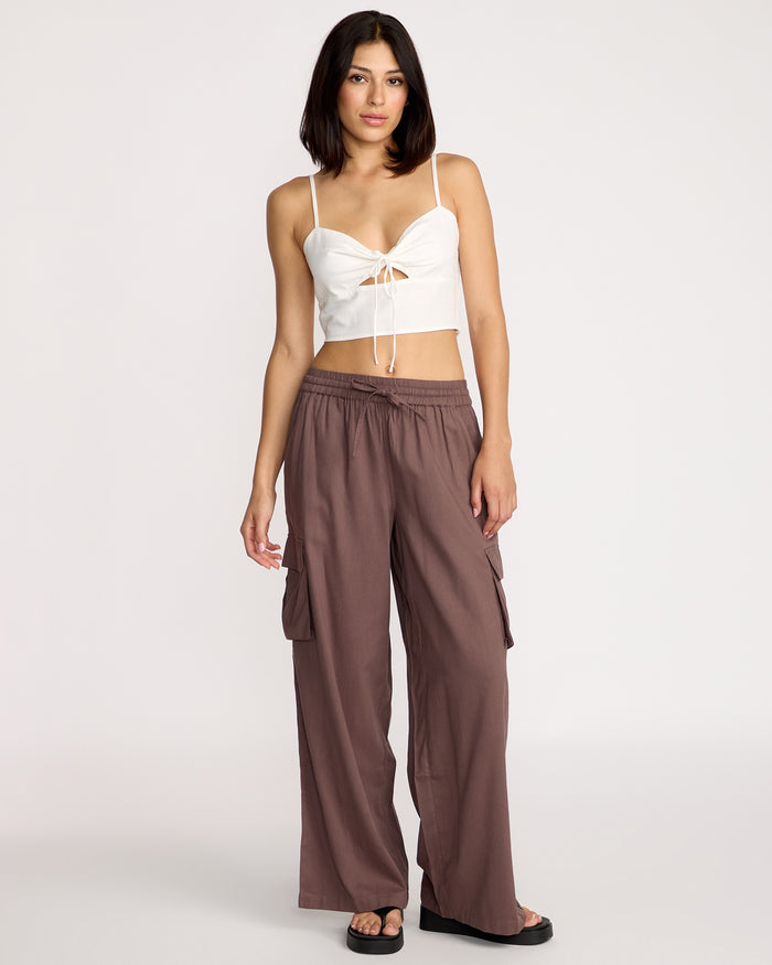 A woman with shoulder-length dark hair stands in a studio against a plain light background, wearing the Volcom Women's Money Mama Cami—a white crop top featuring a smocked back and tied front—paired with loose mauve cargo pants and black sandals.