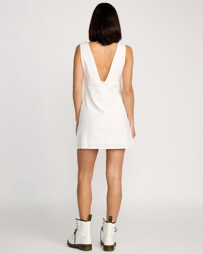 A woman with straight, shoulder-length dark hair is seen from behind in a plain setting, wearing the Volcom Women's Money Mama Dress—a sleeveless short white dress with a deep V-cut back—paired with white ankle-high lace-up boots.
