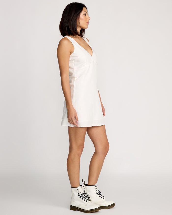 A woman with straight shoulder-length dark hair stands in profile against a plain background, wearing the Volcom Women's Money Mama Dress, a sleeveless white V-neck mini dress, paired with white lace-up ankle boots.