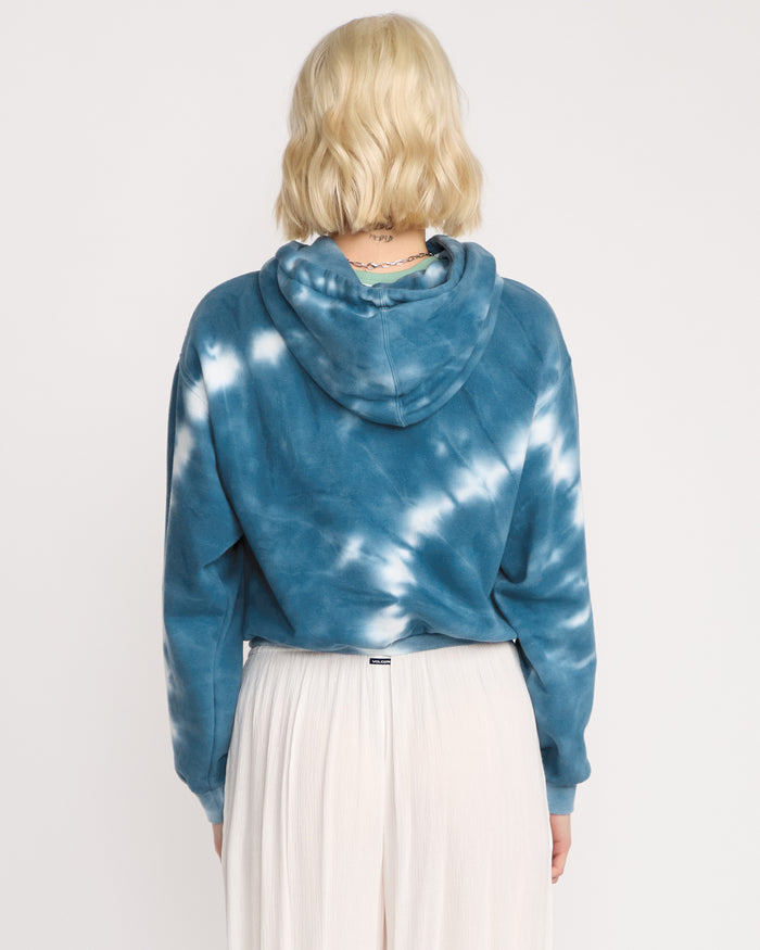 A woman with short blonde hair, seen from behind, wears the Volcom Women's New Tie Dye Hoodie in blue and white paired with a long white pleated skirt, standing against a plain white background.