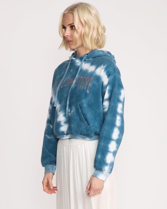 A woman with short blonde hair models the Volcom Women's New Tie Dye Hoodie, featuring blue and white tones and front text, paired with a long white pleated skirt. She stands sideways against a plain white background.