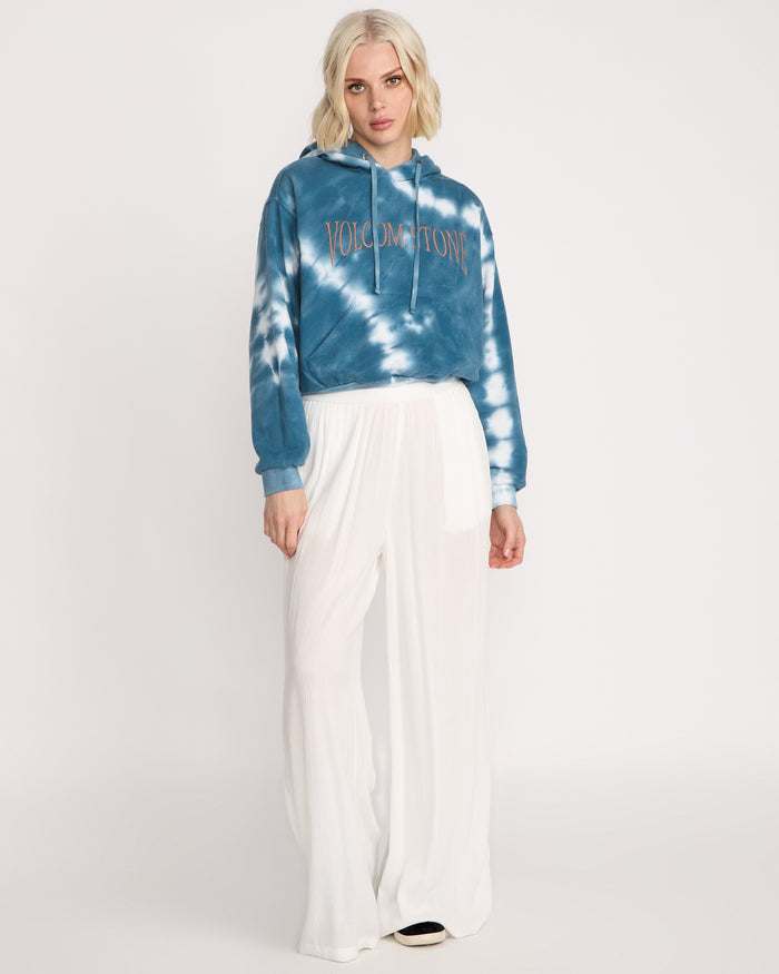 A woman with short blonde hair wears the Volcom Women's New Tie Dye Hoodie and loose white pants, standing against a plain white background.