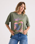 Wearing the Volcom Women's Night Shade Bad Tee—a relaxed, oversized green graphic shirt featuring a vibrant pink and blue tropical print—a woman stands against a plain background with one hand in her jean pocket, gazing confidently at the camera.