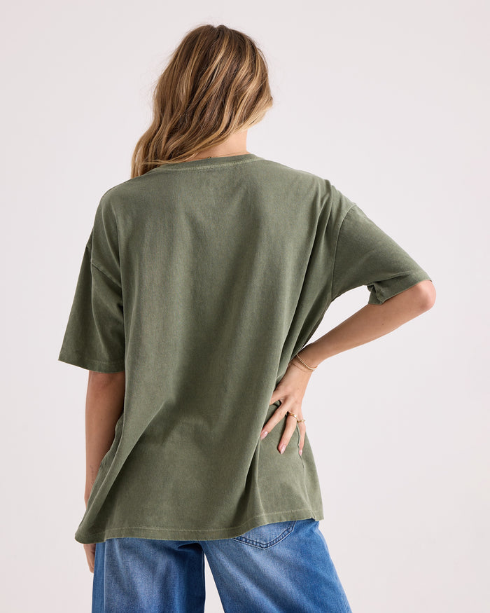 A person with long, wavy hair is seen from behind, wearing the Volcom Women's Night Shade Bad Tee—an oversized 100% cotton olive green graphic T-shirt—paired with blue jeans, one hand resting on their hip against a plain light background.
