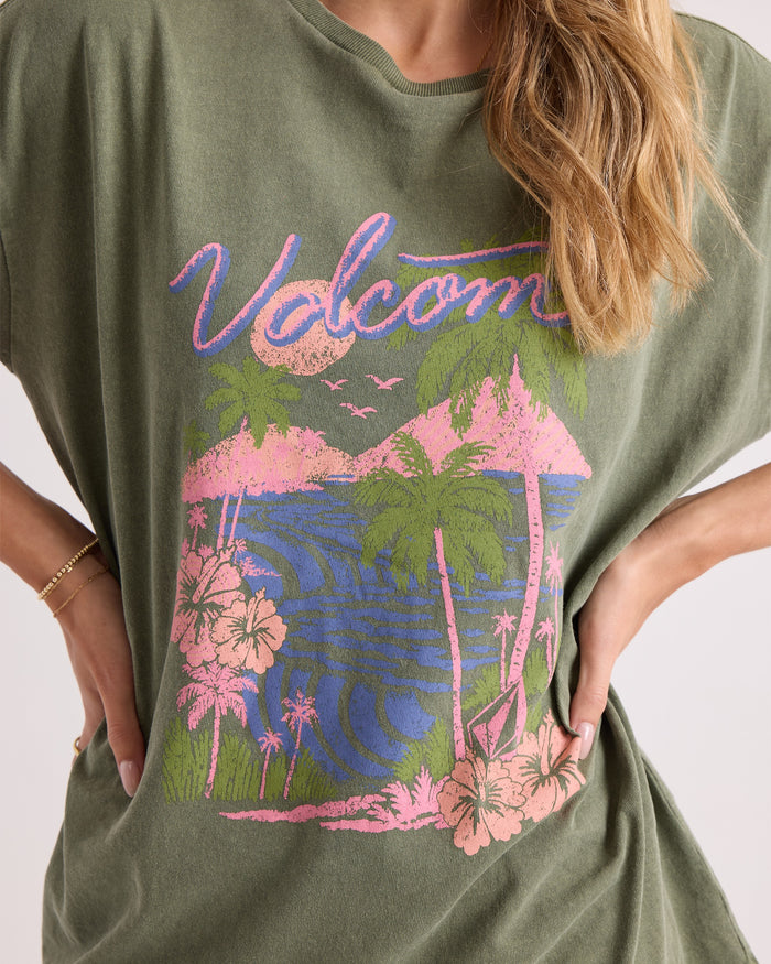 A person wearing the Volcom Women's Night Shade Bad Tee, an oversized 100% cotton green shirt with a pink and blue tropical beach graphic featuring palm trees, flowers, and waves.