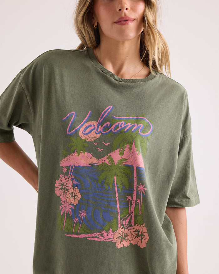 A woman wears the Volcom Women's Night Shade Bad Tee in olive green, featuring a pastel tropical graphic of palm trees, flowers, and water. Made from 100% cotton with short sleeves and a relaxed fit, this tee offers all-day comfort.