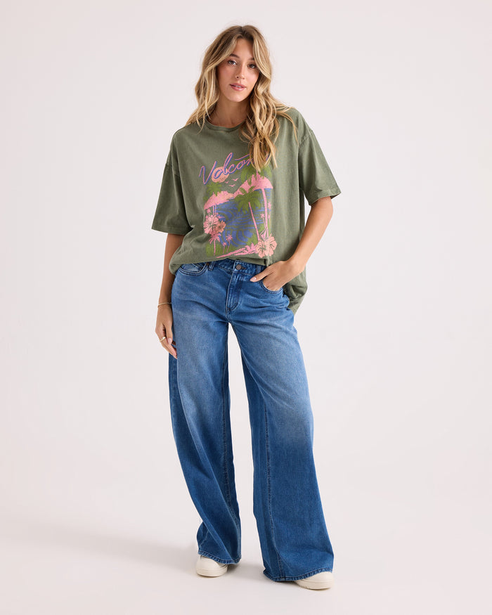 A woman poses casually with one hand in her pocket against a light background, wearing the Volcom Women's Night Shade Bad Tee—an oversized green graphic T-shirt made from 100% cotton—paired with wide-leg blue jeans and white shoes. She has long, wavy hair.