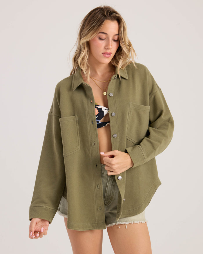 A woman wearing the Volcom Women's Nollie Jacket—an oversized 100% cotton shirt jacket—over a patterned bikini top and light green shorts stands against a plain background, looking down as she adjusts her shirt.